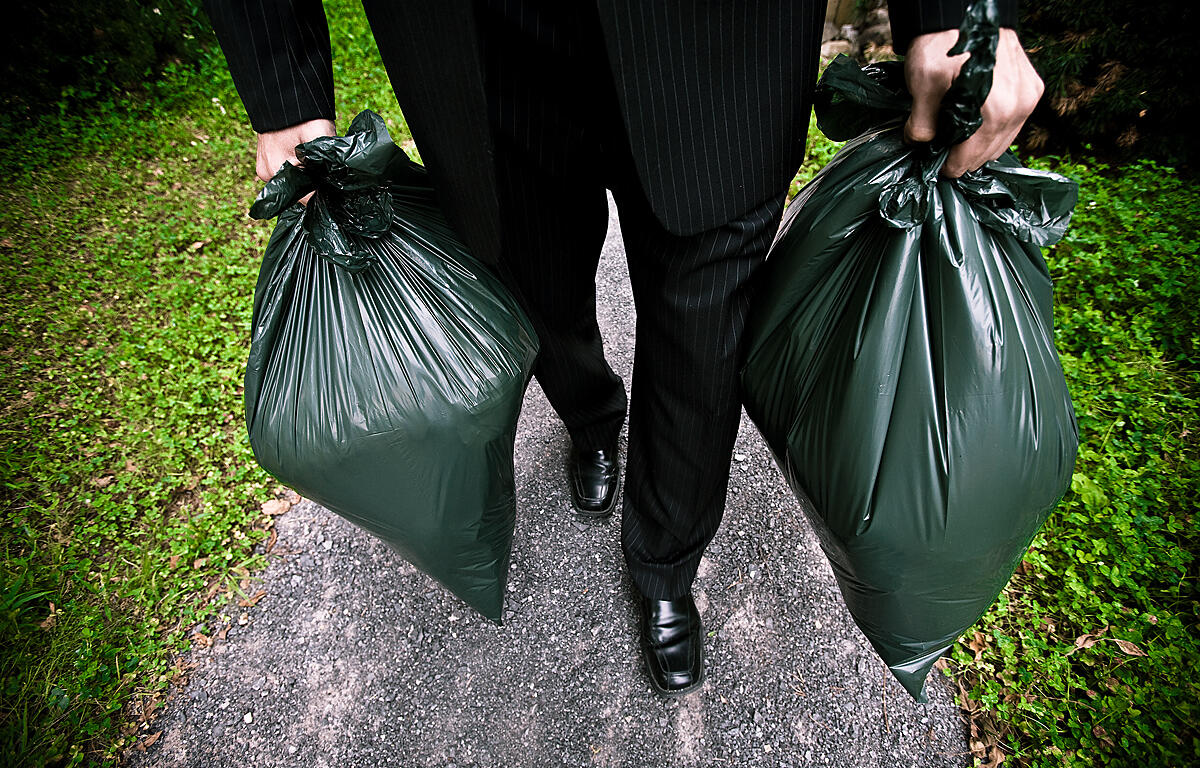 A person in all black carrying a green trash bag in each hand.