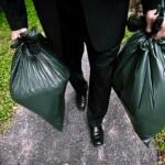 A person in all black carrying a green trash bag in each hand.