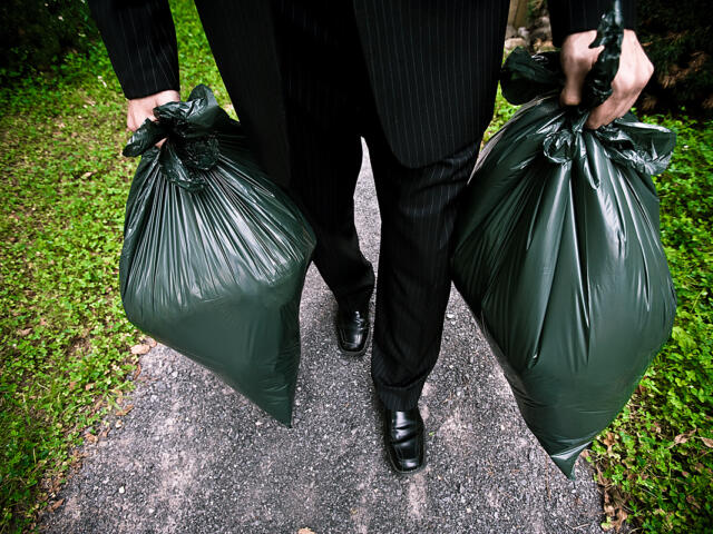 A person in all black carrying a green trash bag in each hand.