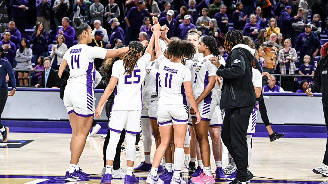 James Madison University women's basketball players huddle in a circle and throw their hands up.