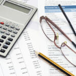 A calculator, glasses, and pencil lay on top of several scattered papers.