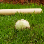 Closeup horizontal photo of old baseball in front of wooden bat on natural grass field