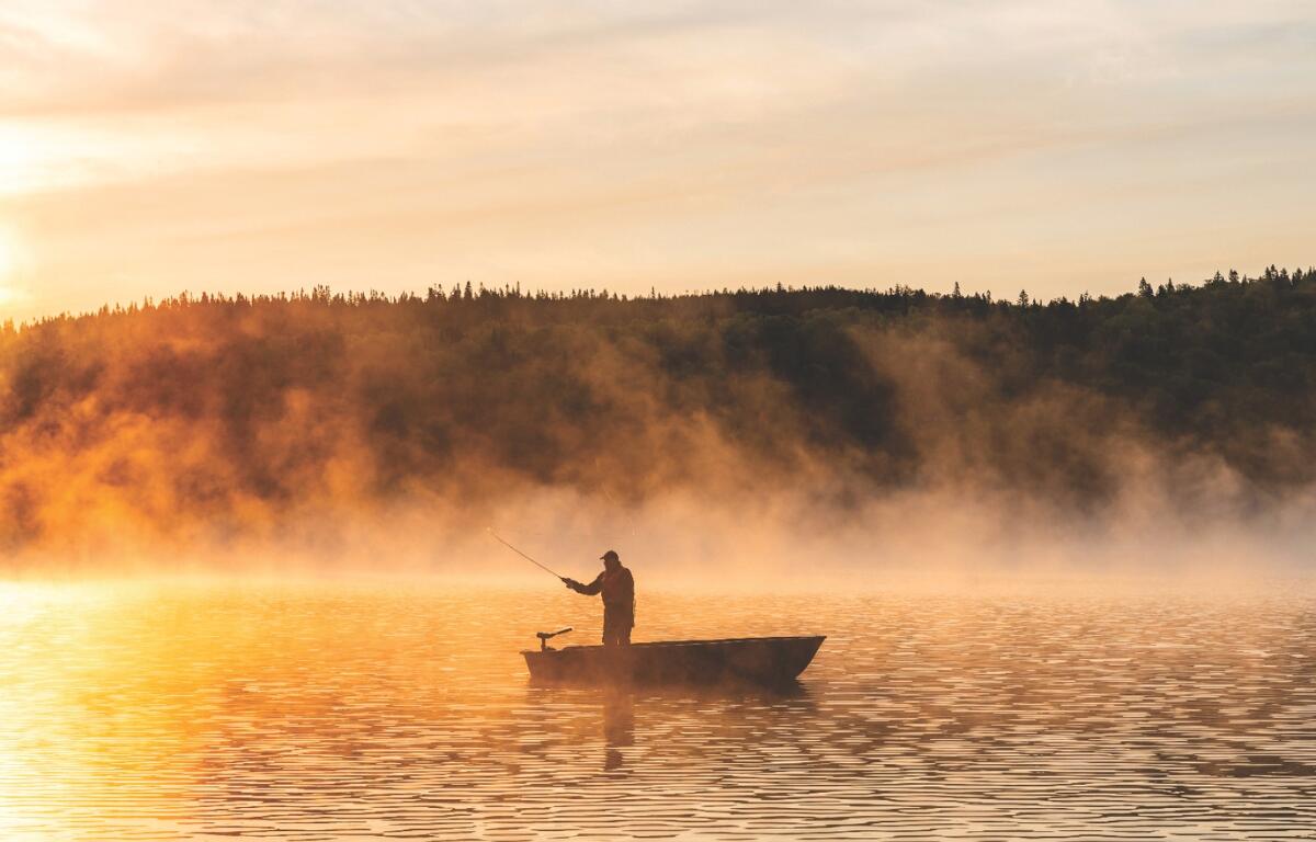 A man fishing on a boat in a lake while the sun rises over the water.