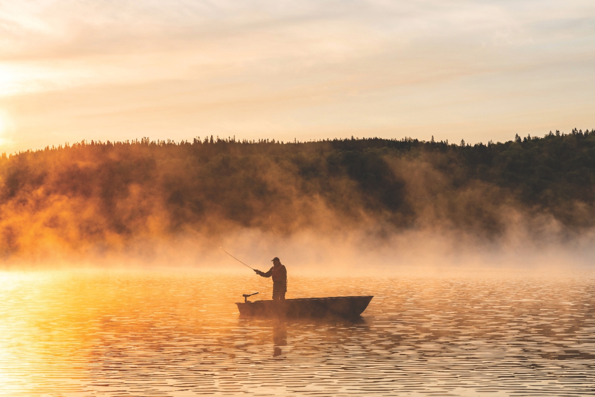 A man fishing on a boat in a lake while the sun rises over the water.