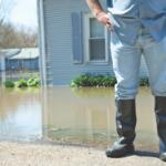 A person in rain boots standing in front of a flooded lawn.