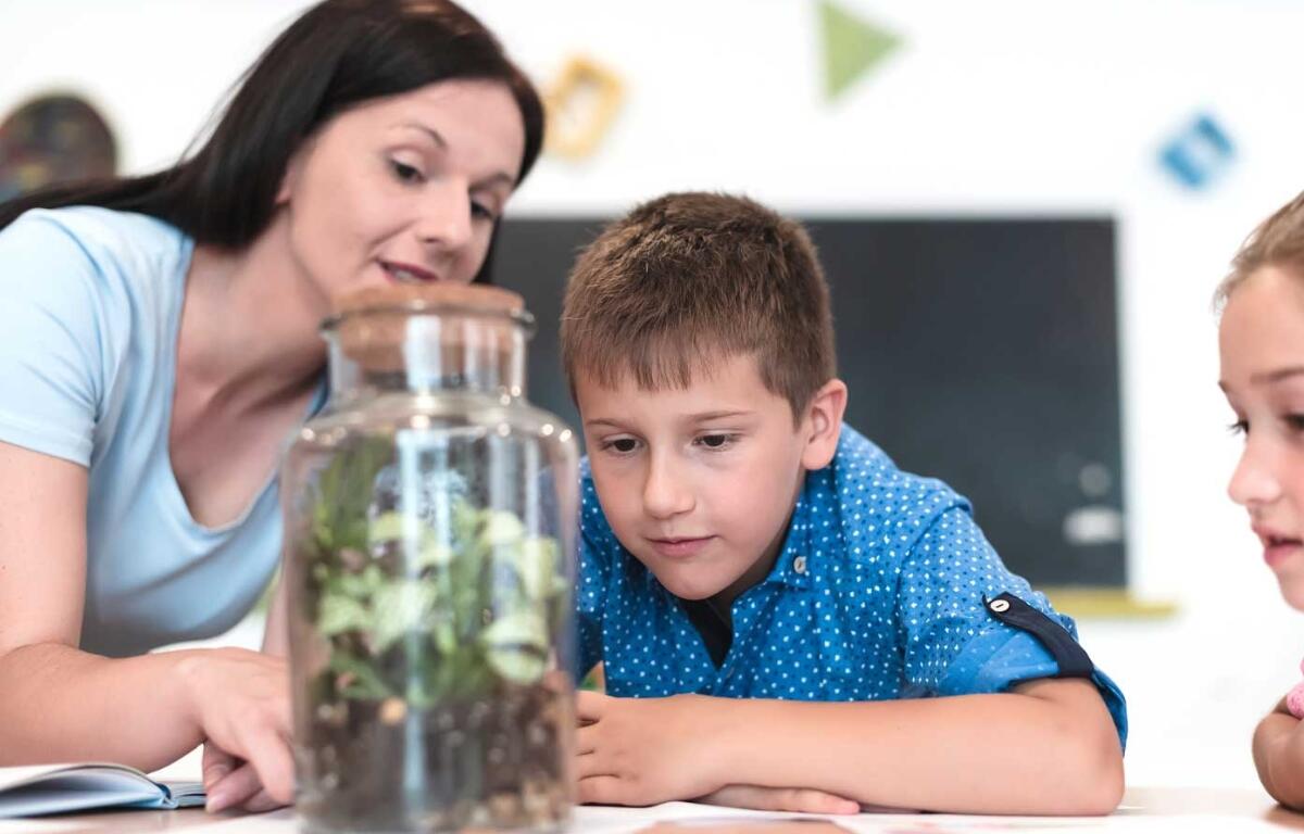 A teacher and student observe plants in a glass jar.