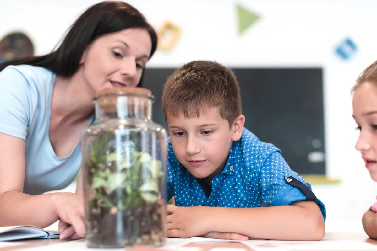 A teacher and student observe plants in a glass jar.