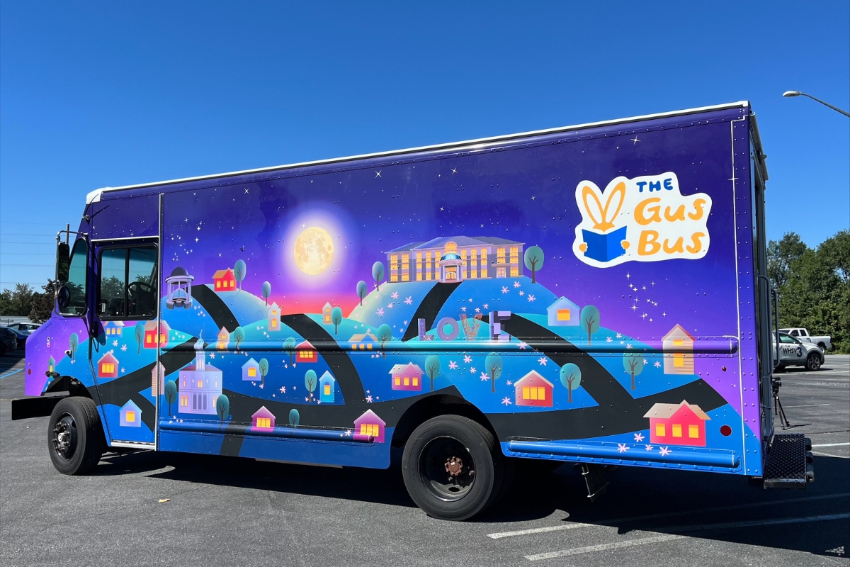 A look at the outside of the new electric Gus Bus. It features a cartoon night scene with a neighborhood and iconic James Madison University buildings under a full moon.