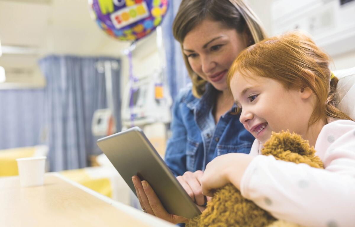 A child in a hospital bed with a stuffed animal and tablet, smiling with a nurse.