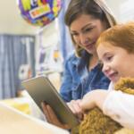 A child in a hospital bed with a stuffed animal and tablet, smiling with a nurse.