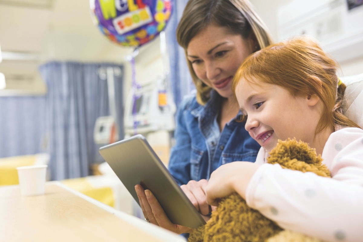 A child in a hospital bed with a stuffed animal and tablet, smiling with a nurse.