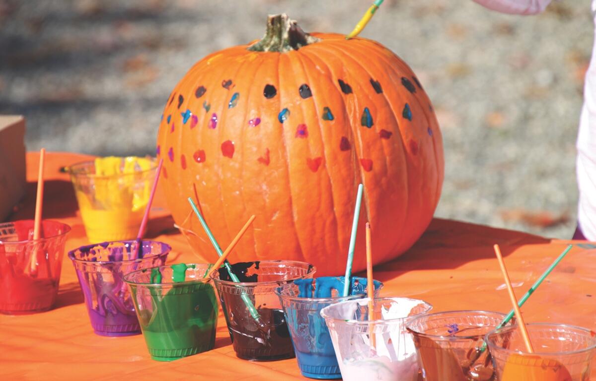 A pumpkin with painted dots on it sits on a table with different cups of paint.