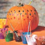 A pumpkin with painted dots on it sits on a table with different cups of paint.