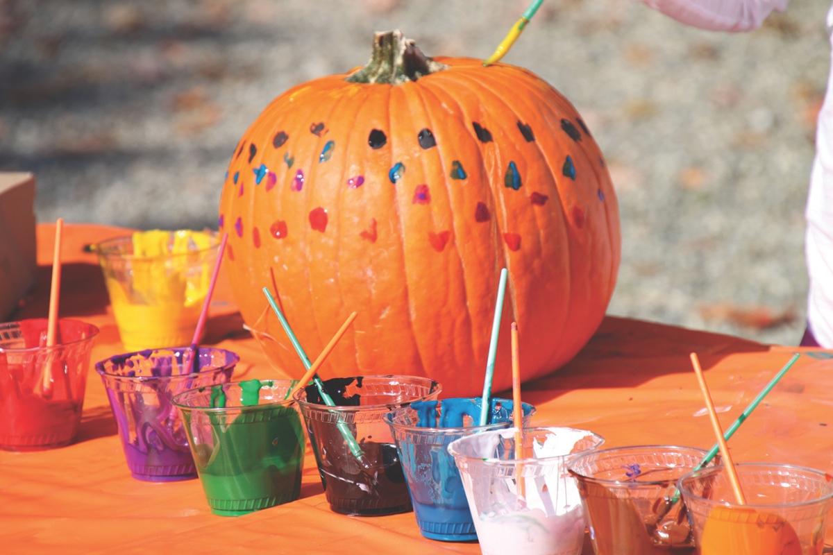 A pumpkin with painted dots on it sits on a table with different cups of paint.