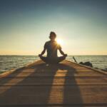 A person meditating on a pier near water.