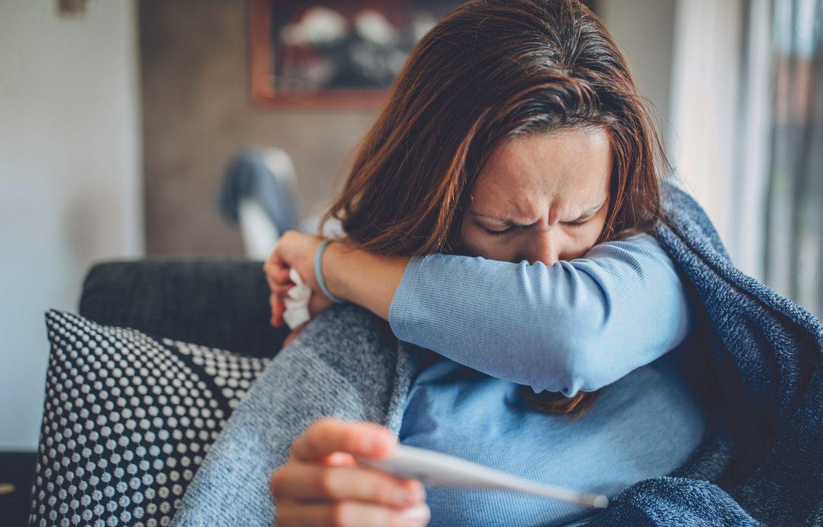 Woman coughing into her arm with a thermometer in the other hand