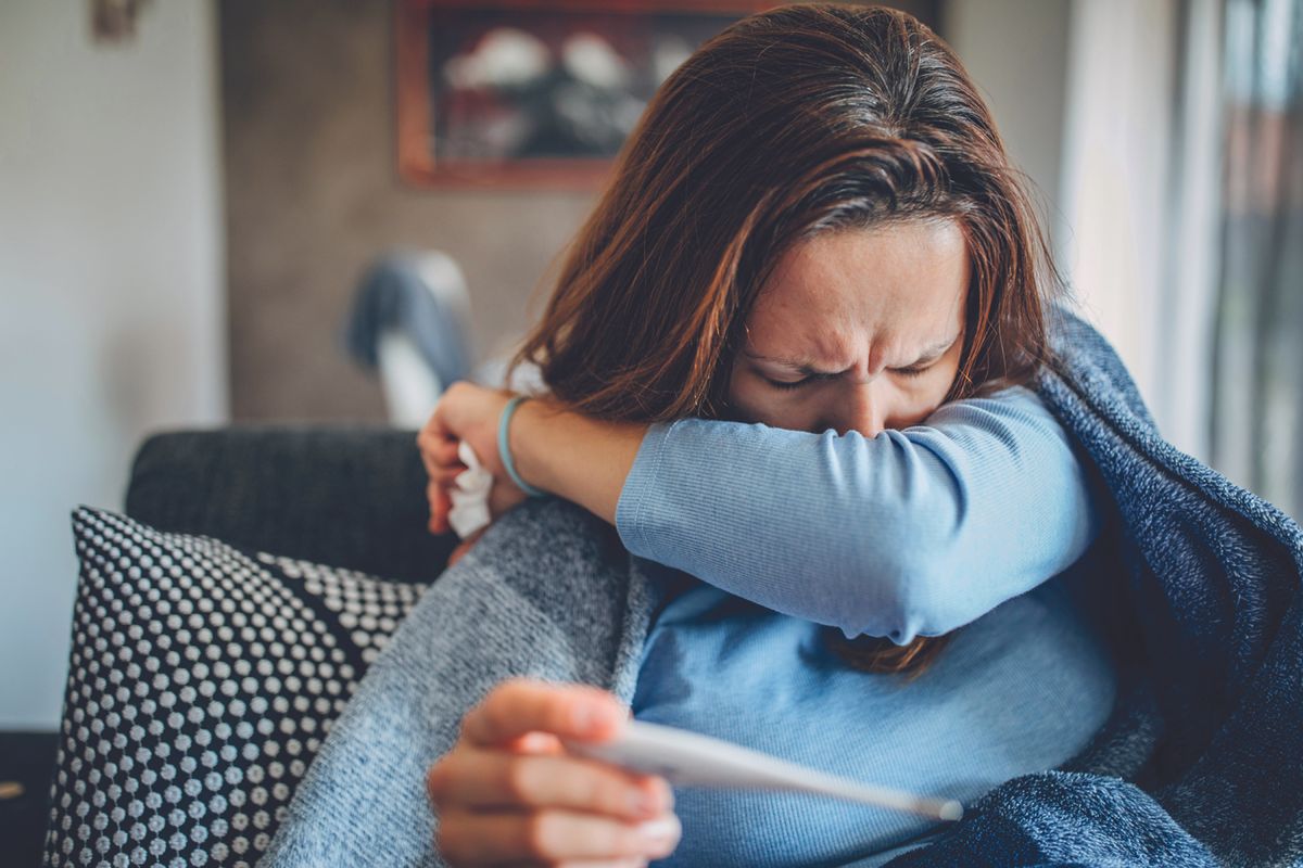 Woman coughing into her arm with a thermometer in the other hand