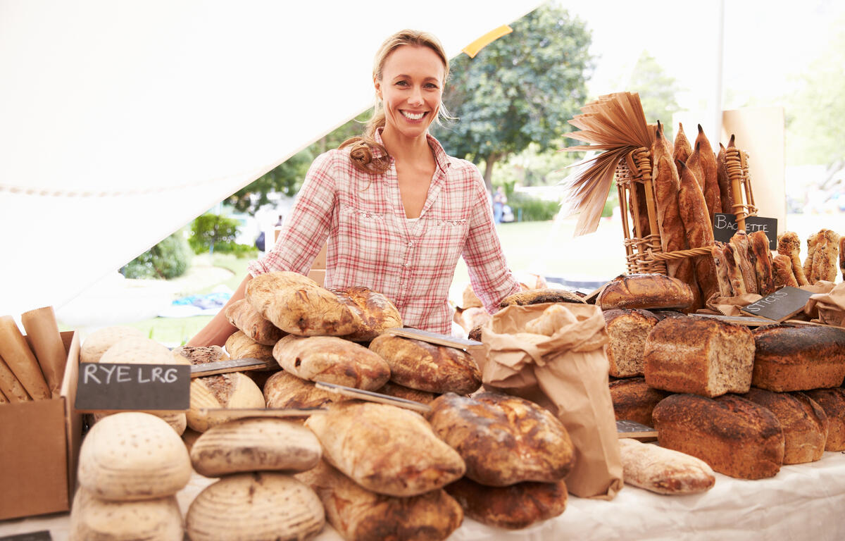 Smiling vendor at an outdoor bread stall with loaves and baguettes on display.