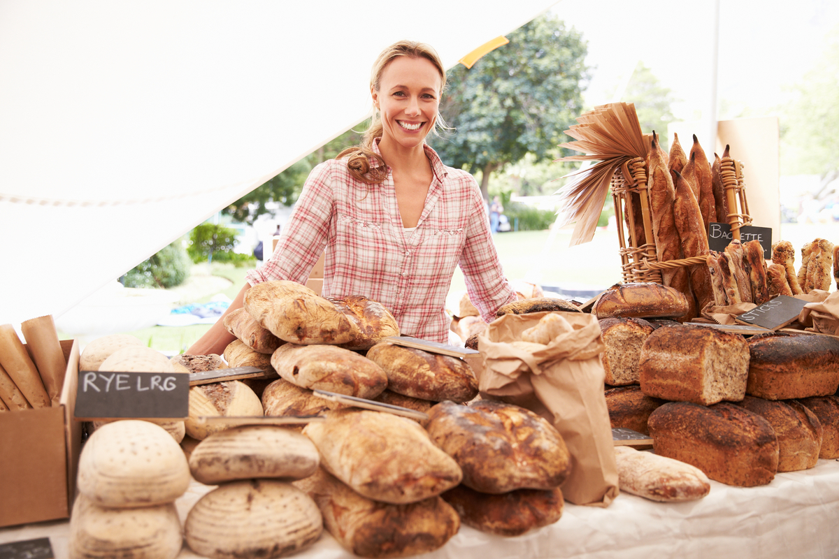 Smiling vendor at an outdoor bread stall with loaves and baguettes on display.
