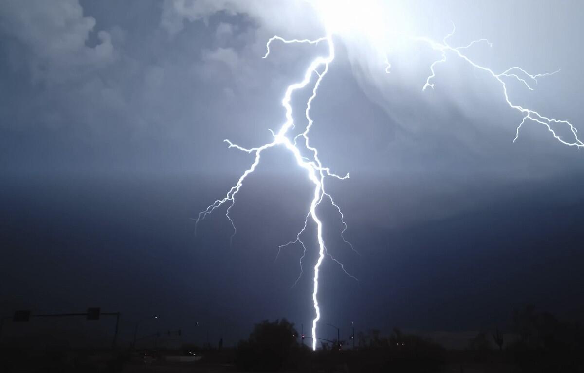 Bright lightning bolt striking from a stormy night sky onto a dark urban landscape below, streetlights silhouetted.