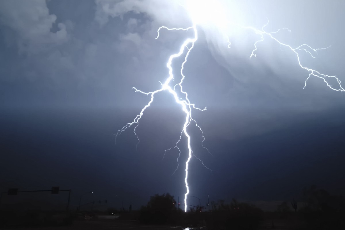 Bright lightning bolt striking from a stormy night sky onto a dark urban landscape below, streetlights silhouetted.