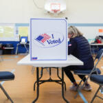 Voter seated at a table in a gym, casting a ballot at a privacy booth marked 'I Voted'.