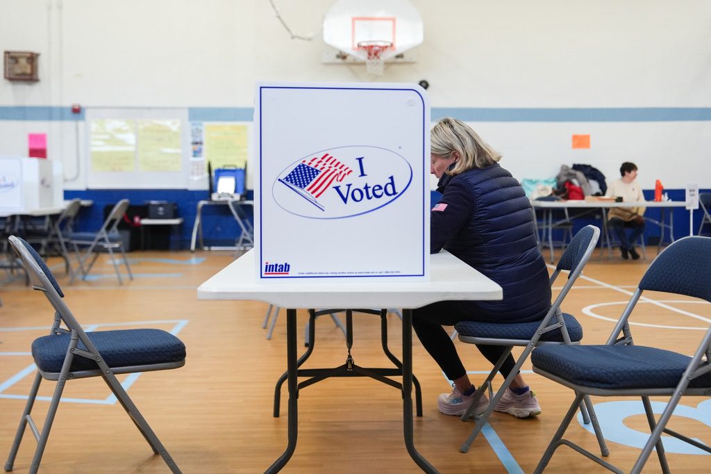 Voter seated at a table in a gym, casting a ballot at a privacy booth marked 'I Voted'.