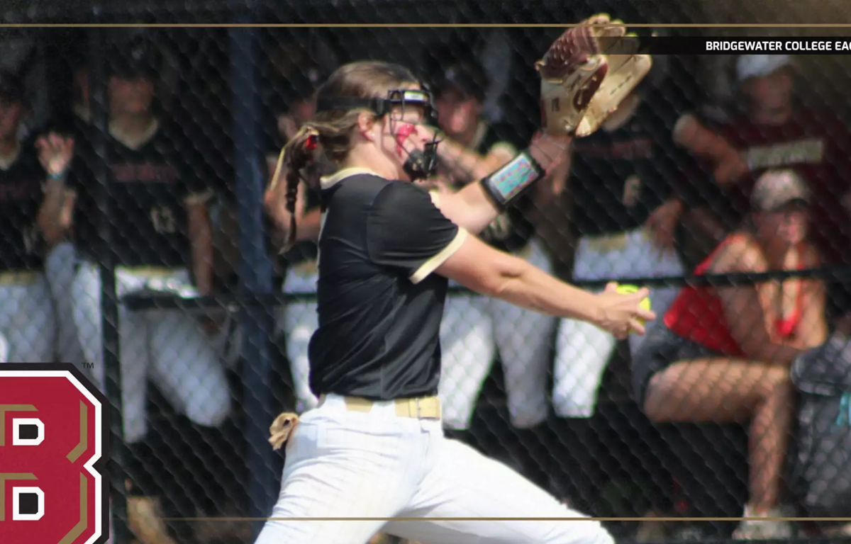 Female softball pitcher in a black shirt and white pants mid-pitch, with fans behind a chain-link fence.