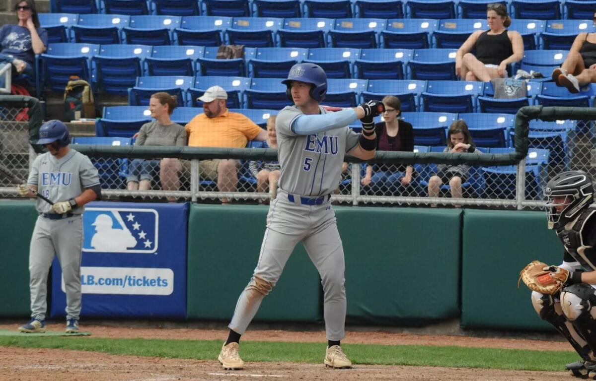 Baseball batter in EMU gray uniform swinging at a pitch, number 5, wearing helmet; catcher in black gear crouches to the right; spectators in blue seats behind.