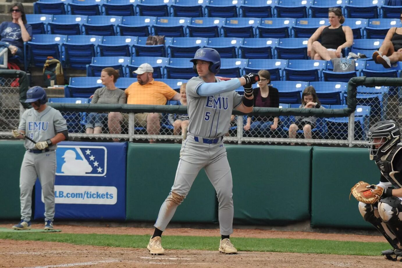 Baseball batter in EMU gray uniform swinging at a pitch, number 5, wearing helmet; catcher in black gear crouches to the right; spectators in blue seats behind.