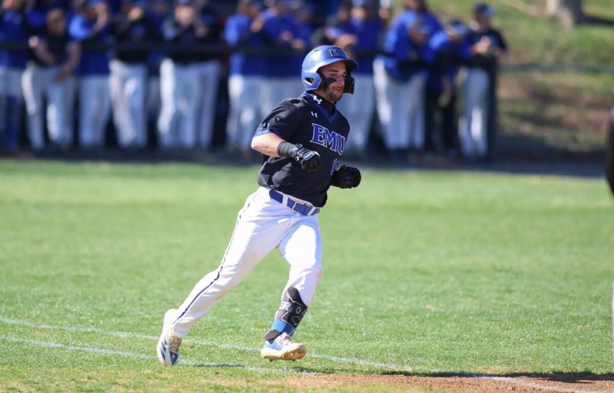 Baseball player in a dark EMU jersey running on the field, wearing a helmet and white pants with a crowd in the background. 20-23? Wait no.