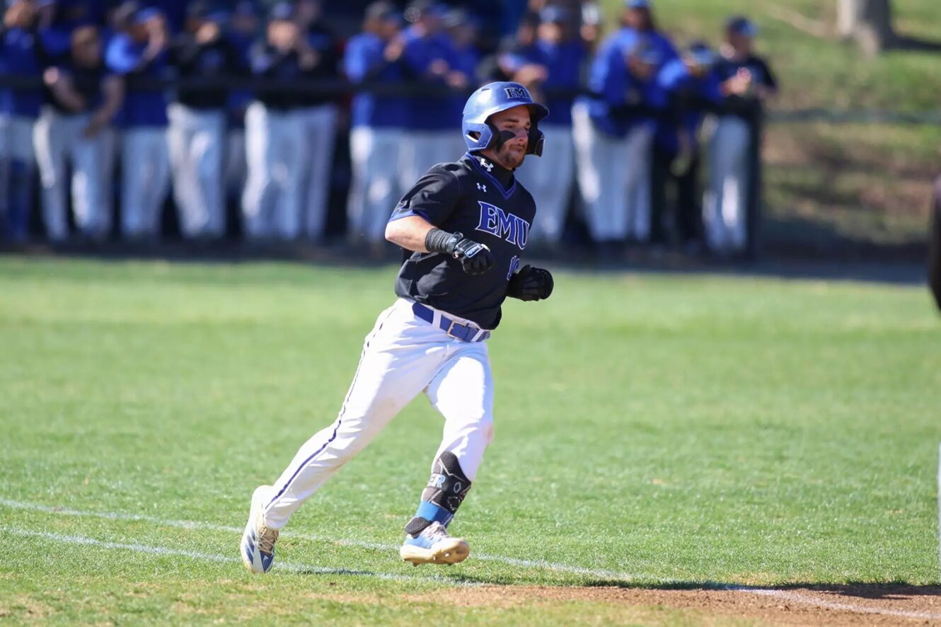 Baseball player in a dark EMU jersey running on the field, wearing a helmet and white pants with a crowd in the background. 20-23? Wait no.