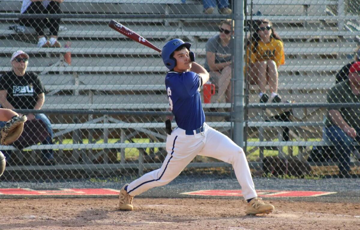 Baseball batter in a blue uniform swings a bat during a game, wearing a helmet with spectators behind a chain-link fence in the stands.