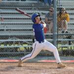 Baseball batter in a blue uniform swings a bat during a game, wearing a helmet with spectators behind a chain-link fence in the stands.