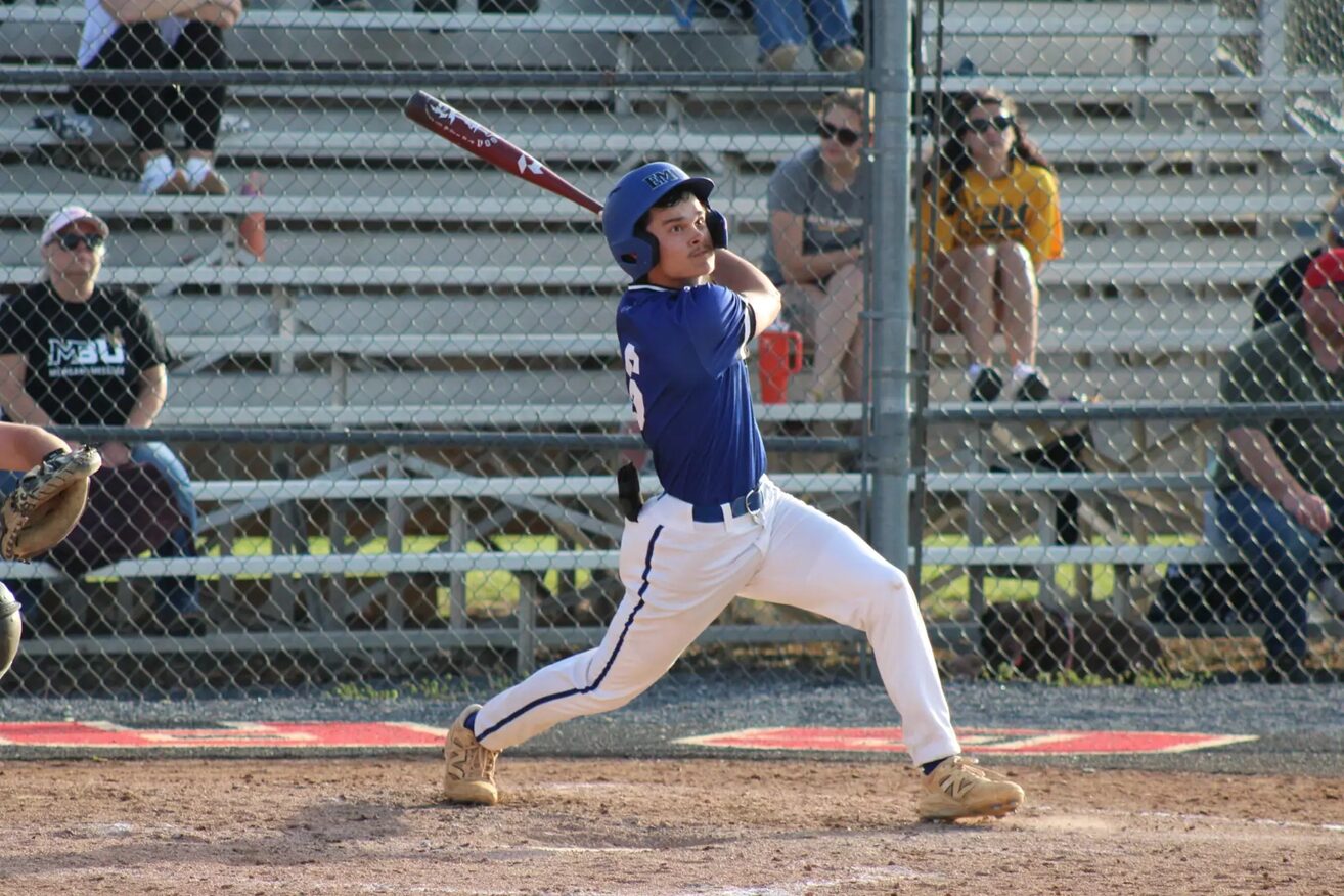 Baseball batter in a blue uniform swings a bat during a game, wearing a helmet with spectators behind a chain-link fence in the stands.