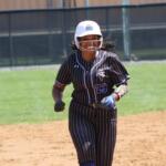 Female softball player in a pinstriped black uniform runs on the infield, wearing a white helmet and blue batting gloves.