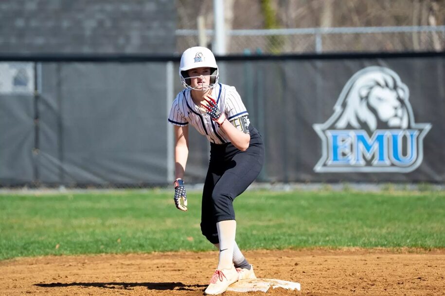 Female softball player in a striped uniform and helmet on a dirt infield, standing on a base with a glove, ready to throw or field; EMU logo on the outfield fence in the background.