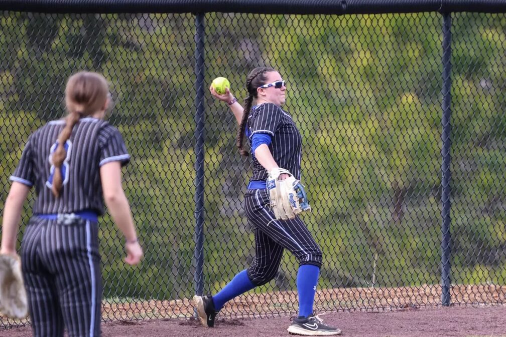 Softball players in pinstripe uniforms on a diamond; one pitcher throws a ball with glove ready, others watch behind a chain-link fence.