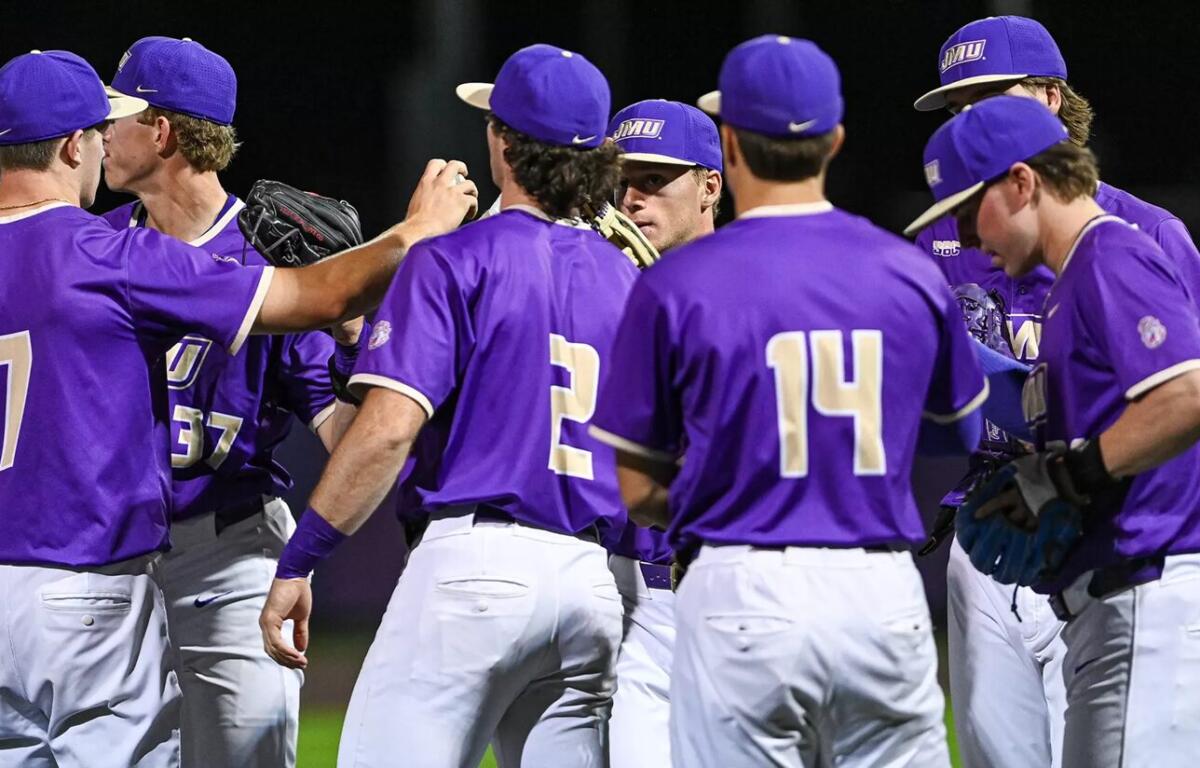 A group of baseball players in purple uniforms huddle on the field during a night game, backs to the camera with visible numbers 7, 2, and 14.