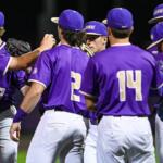 A group of baseball players in purple uniforms huddle on the field during a night game, backs to the camera with visible numbers 7, 2, and 14.