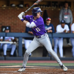 Baseball batter in purple jersey swinging at a pitch, with catcher behind home plate and umpire nearby
