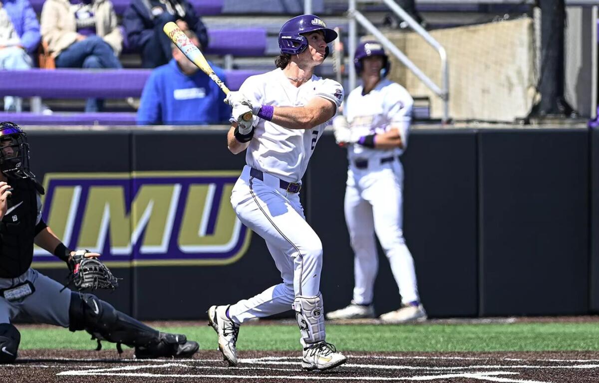 Baseball batter in a white uniform swings at a pitch at home plate, with a catcher crouched behind and purple stadium seating in the background.