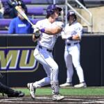 Baseball batter in a white uniform swings at a pitch at home plate, with a catcher crouched behind and purple stadium seating in the background.