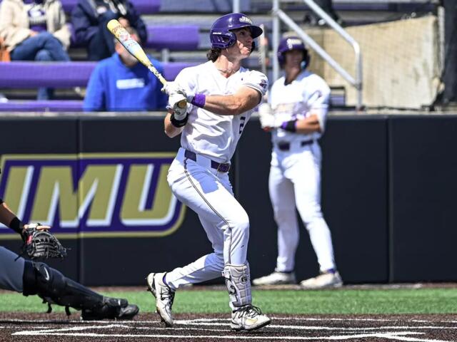 Baseball batter in a white uniform swings at a pitch at home plate, with a catcher crouched behind and purple stadium seating in the background.