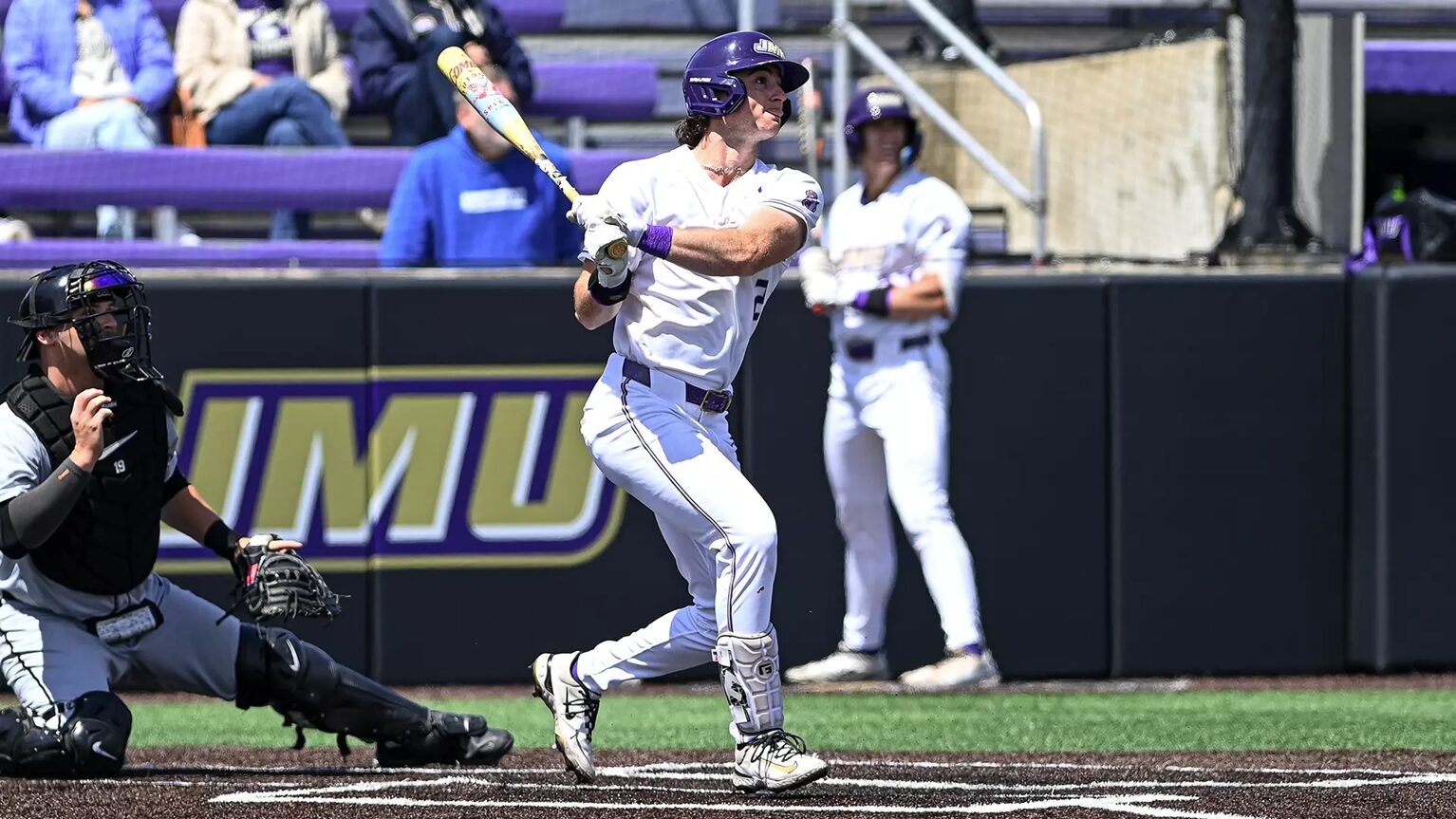 Baseball batter in a white uniform swings at a pitch at home plate, with a catcher crouched behind and purple stadium seating in the background.