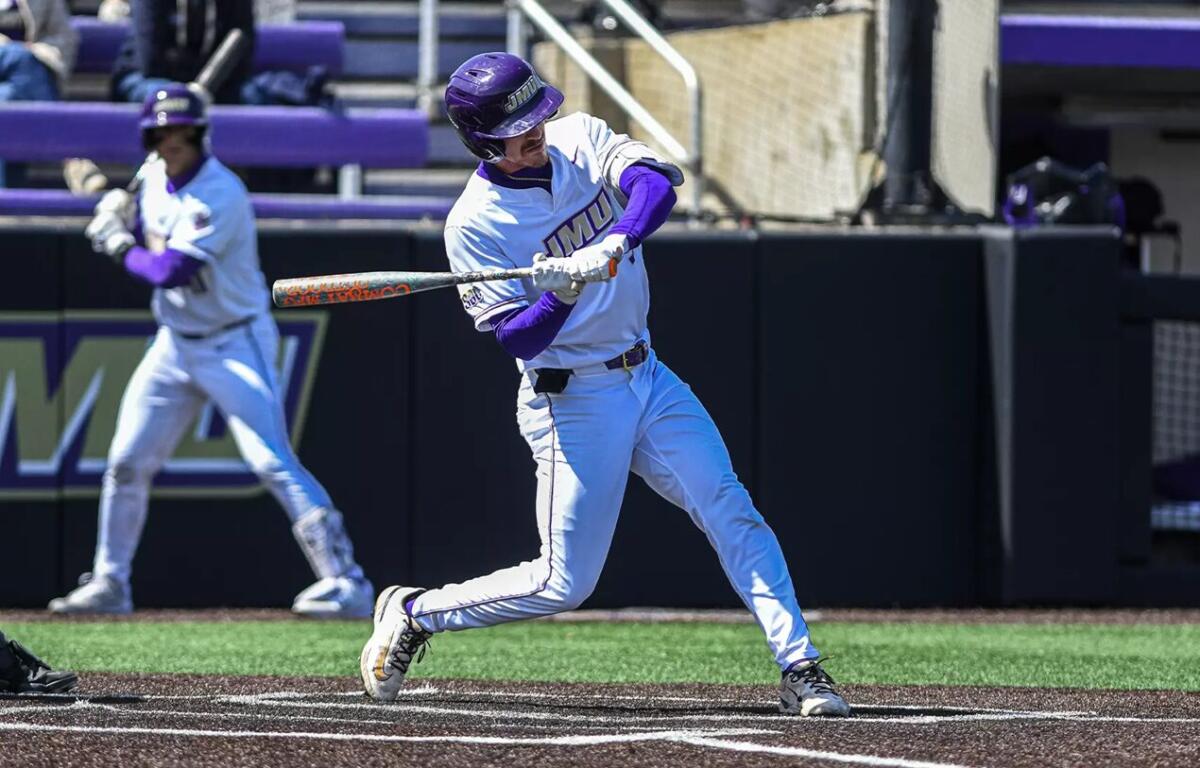 Baseball batter in white and purple uniform swings at a pitch at home plate with a purple helmet visible.