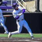Baseball batter in white and purple uniform swings at a pitch at home plate with a purple helmet visible.