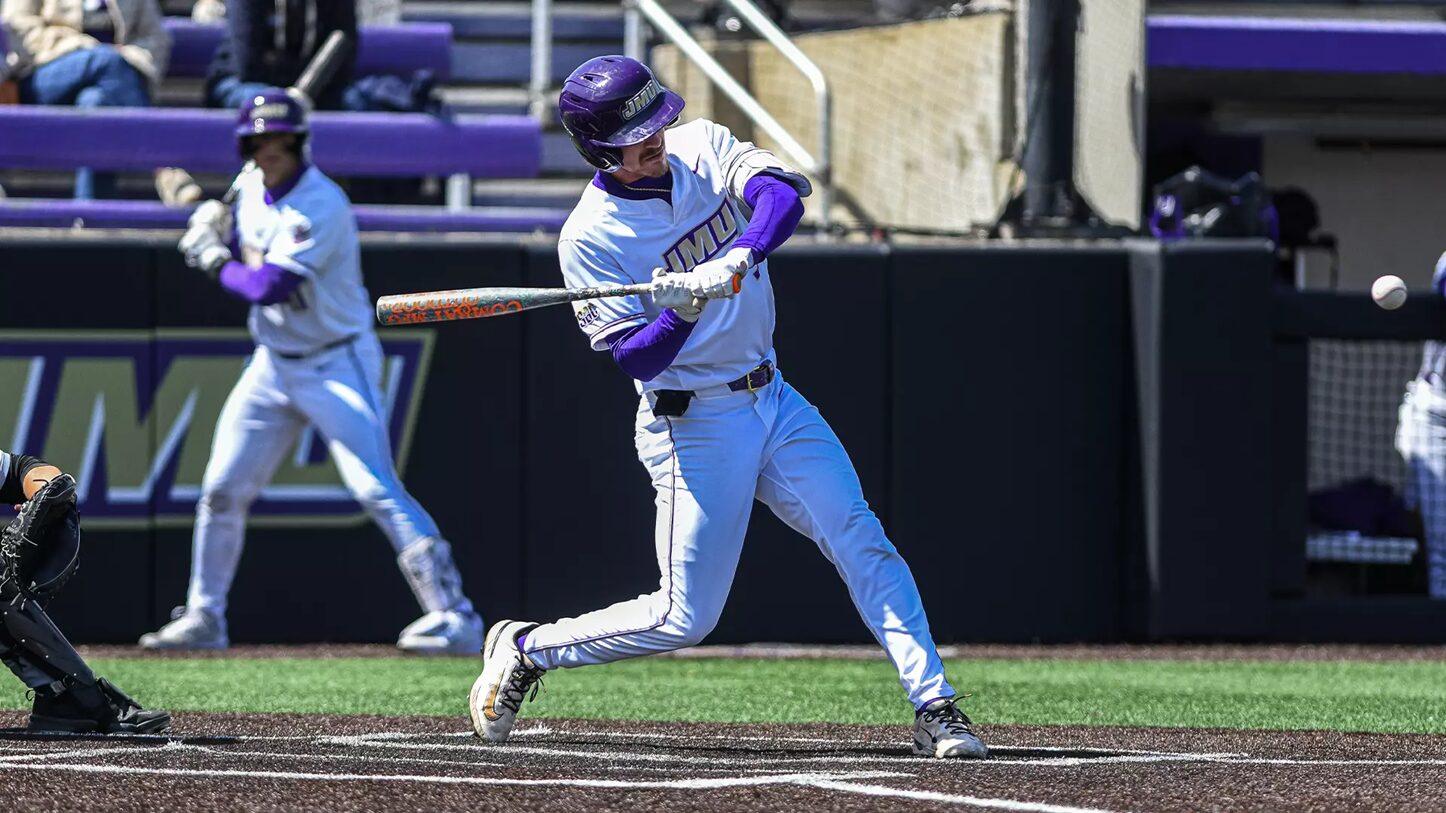 Baseball batter in white and purple uniform swings at a pitch at home plate with a purple helmet visible.