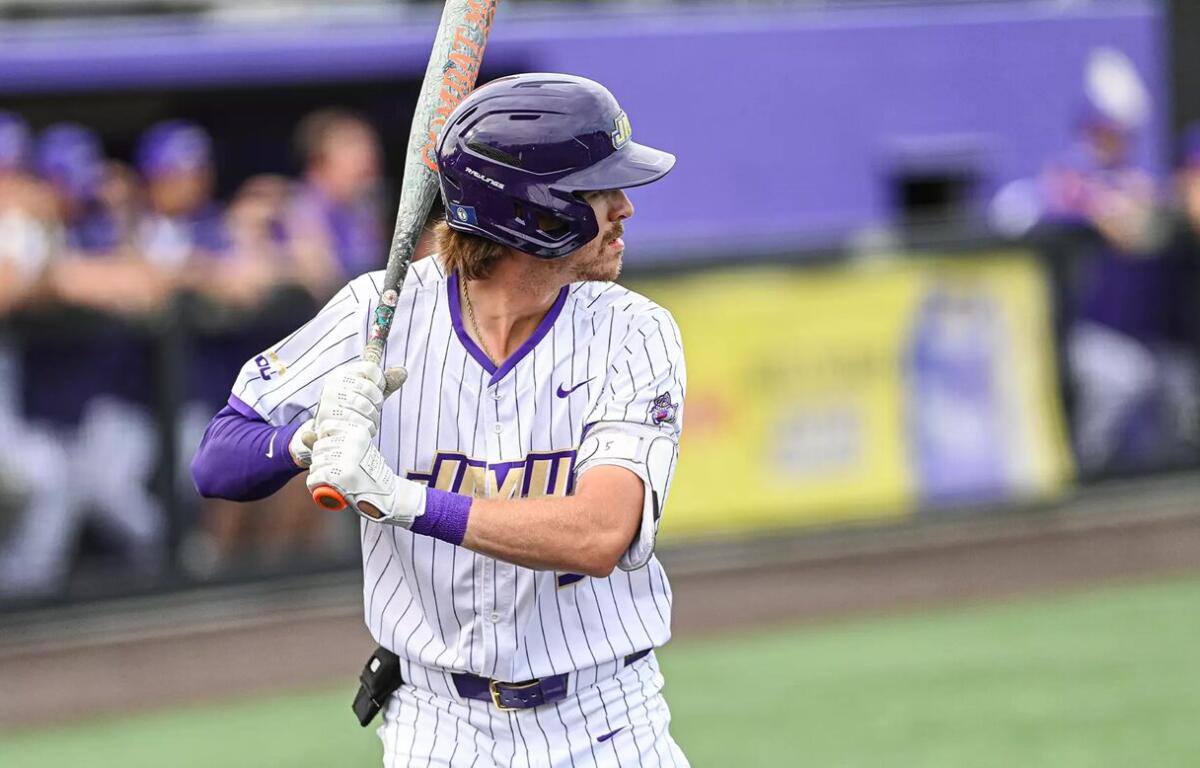 Baseball player in white pinstripe uniform with purple accents, helmet on, mid-swing with bat during a game.