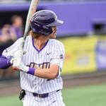 Baseball player in white pinstripe uniform with purple accents, helmet on, mid-swing with bat during a game.